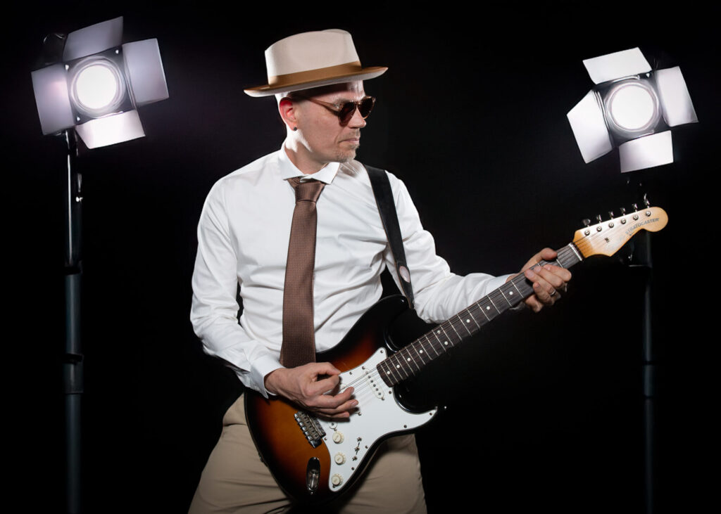 Sandy Leresche à la guitare électrique entre deux projecteurs, sur fond noir